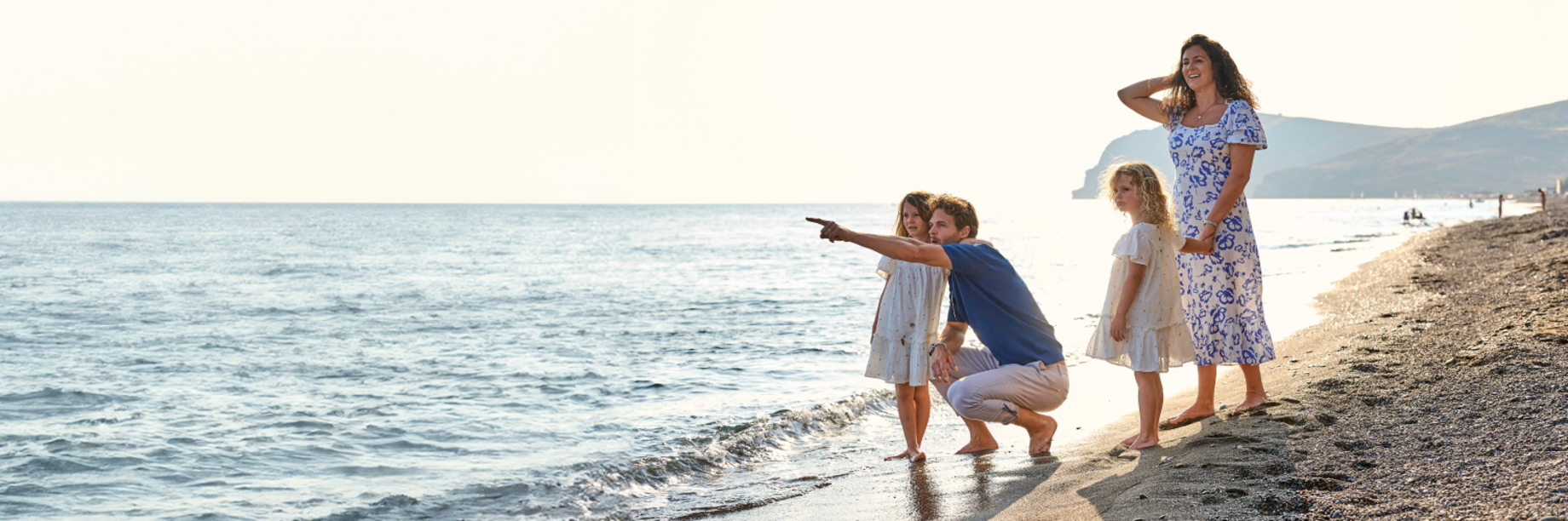 A Family with a mum, dad and 2 small girls, standing on a beach by the sea in Greece