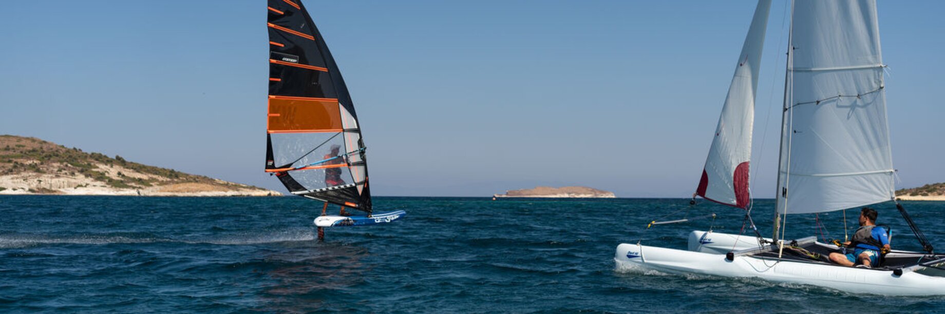 A catamaran sailing alongside a foiling windsurfer