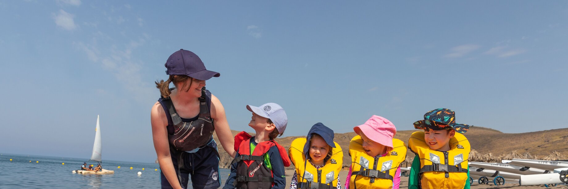 Mark Warner female watersports staff member with a group of young children standing on the beach by a red paddle boarding, all holding an oar together