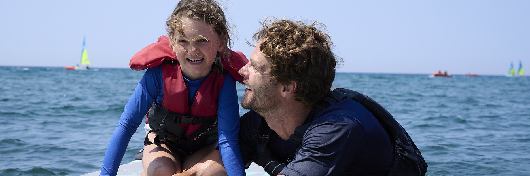 A young girl kneeling on a stand-up paddle board with an instructor in the sea