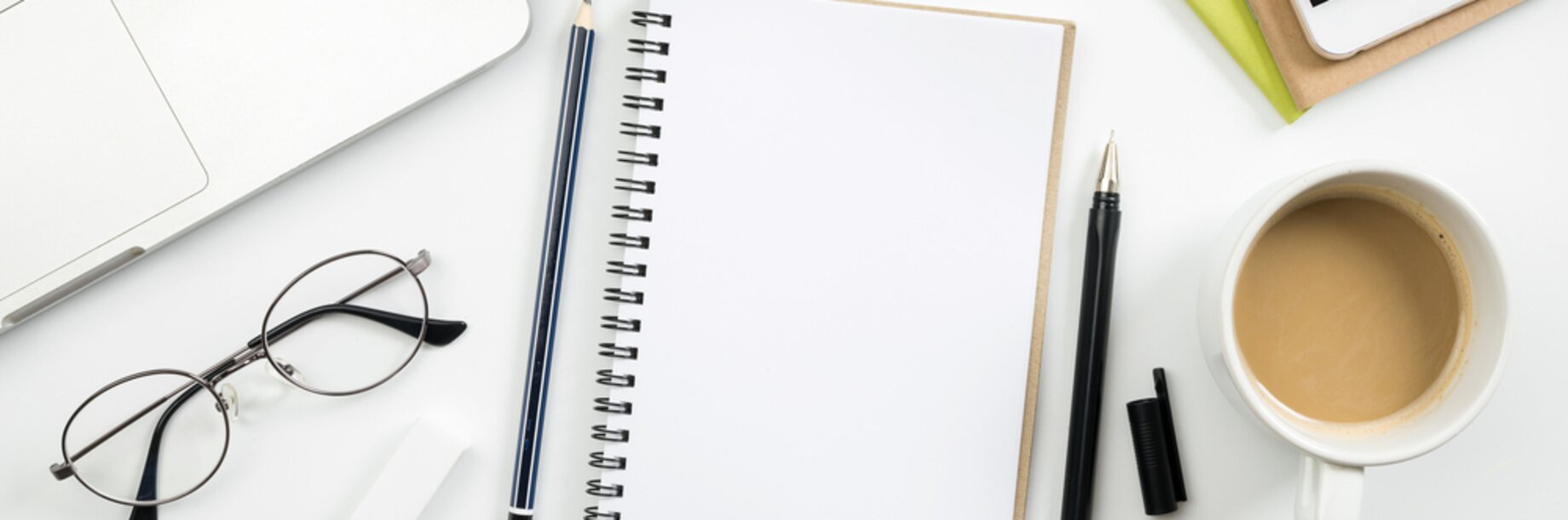 A desk with part of a laptop showing, pad, pen, pencil, mobile phone, glasses, paper clips, small plant and cup of coffee